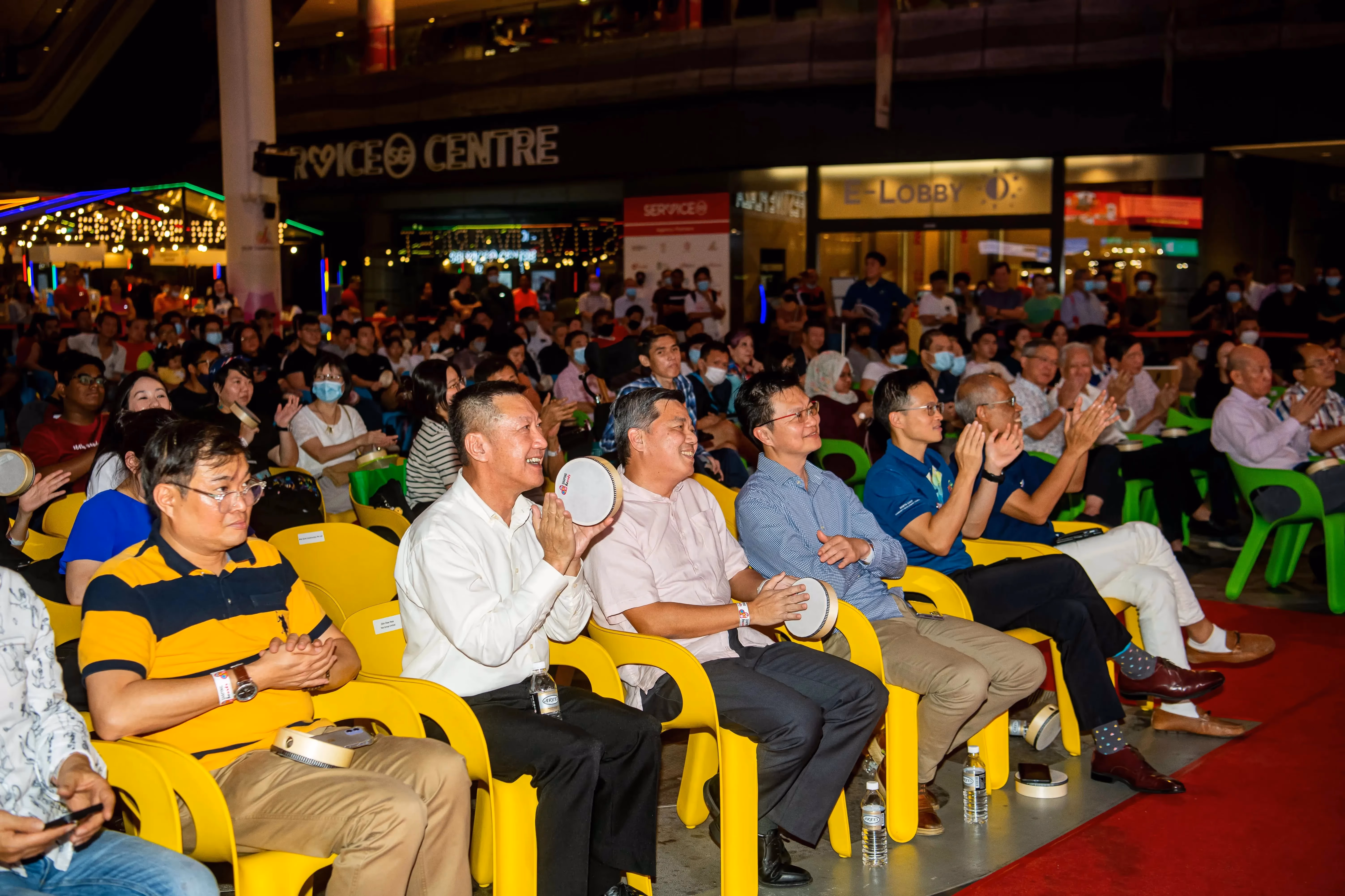 A large group of people is seated in bright yellow chairs, some holding tambourines and clapping in front of a service center.